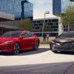 A red 2019 Toyota Camry is parked next to a dark grey one in front of buildings with lots of windows.