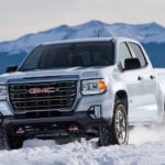 A white 2021 GMC Canyon AT4 is driving through the snow with mountains in the background.