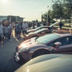 A row of cars and people are shown on a sunny day at a car meet.