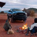 A blue Ram 2500 is shown parked next a family enjoying a campfire.