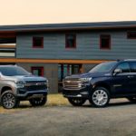 A silver 2023 Chevy Suburban Z71 and a dark blue High Country are shown facing each other in front of a house.