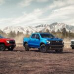 Three 2023 Chevy Silverado 1500s, available at a Chevy truck dealer, are shown parked in the dirt with mountains in the background.