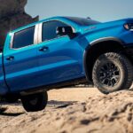 One of the most popular Chevy trucks for sale, a blue 2024 Chevy Silverado 1500 ZR2 is shown driving up a rocky desert road.