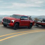 A red 2024 Toyota Tundra for sale is shown towing a boat on a highway.