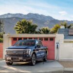 A blue 2022 Ford F-150 Lightning plugged in to charge at a modern home in the desert.