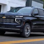 A blue 2025 Chevy Suburban is shown driving on a highway to visit a Fayetteville Chevy Dealer.