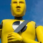 A yellow crash dummy is shown holding his seatbelt near a used car dealer.