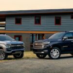 A silver 2024 Chevy Suburban Z71 and a blue 2024 Chevy Suburban High Country parked in front of a house.