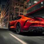 An orange 2024 Toyota GR Supra is shown driving on a city street.