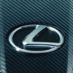 A close-up of the silver Lexus logo mounted on a black carbon fiber grille at a used Lexus dealer.