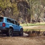 A blue 2024 Toyota Land Cruiser is shown driving off-road near water after visiting a Toyota dealer.