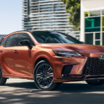 An orange 2024 Lexus RX F SPORT driving past colorful city windows.