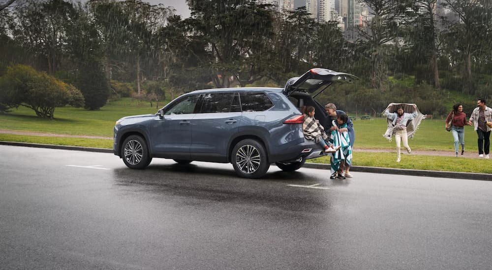 A family is running towards a blue 2024 Toyota Grand Highlander Platinum Hybrid on a rainy day.