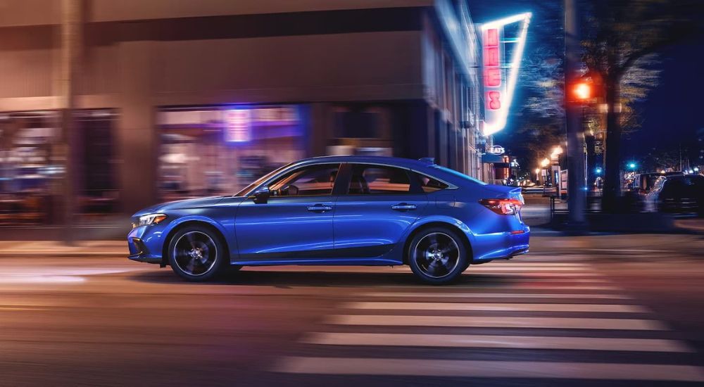 Side angle view of a blue 2025 Honda Civic Hybrid driving in a city from a Honda dealer.