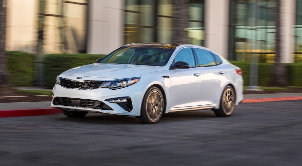 A white 2019 Kia Optima is shown driving on a city street.