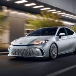 A silver 2025 Toyota Camry for sale is shown driving on a city street.