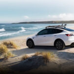 A white 2024 Tesla Model Y is shown parked near the ocean.