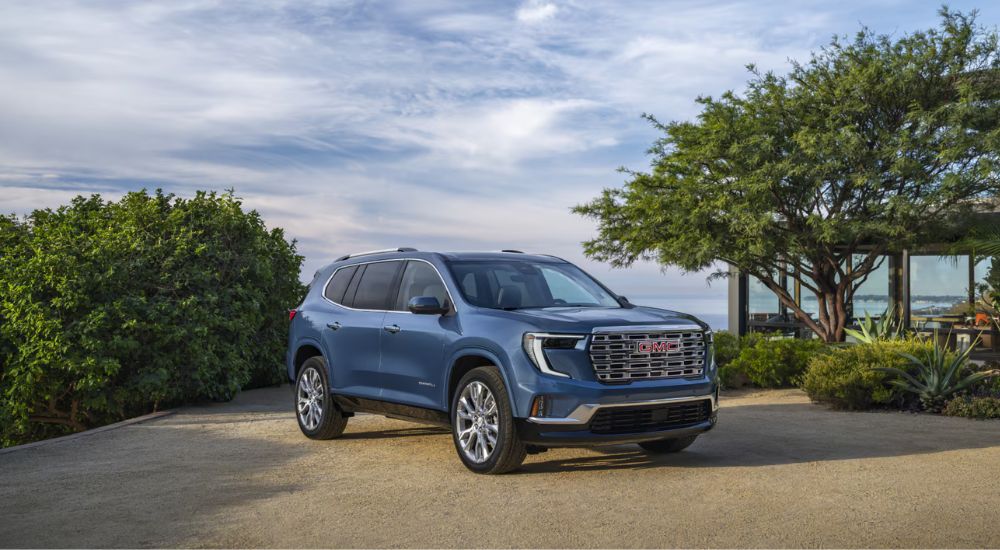 A blue 2025 GMC Acadia Denali parked near trees under a blue sky.