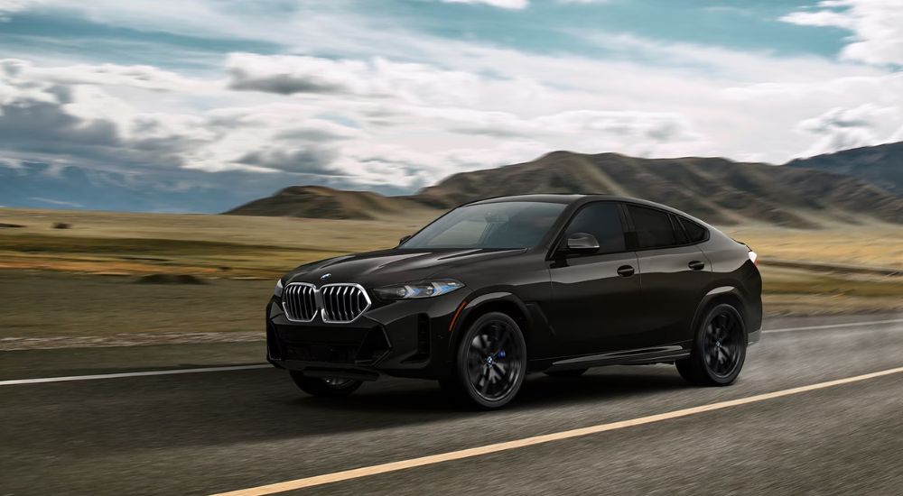 A black 2025 BMW X6 xDrive40i driving on an open road with mountains in the distance.